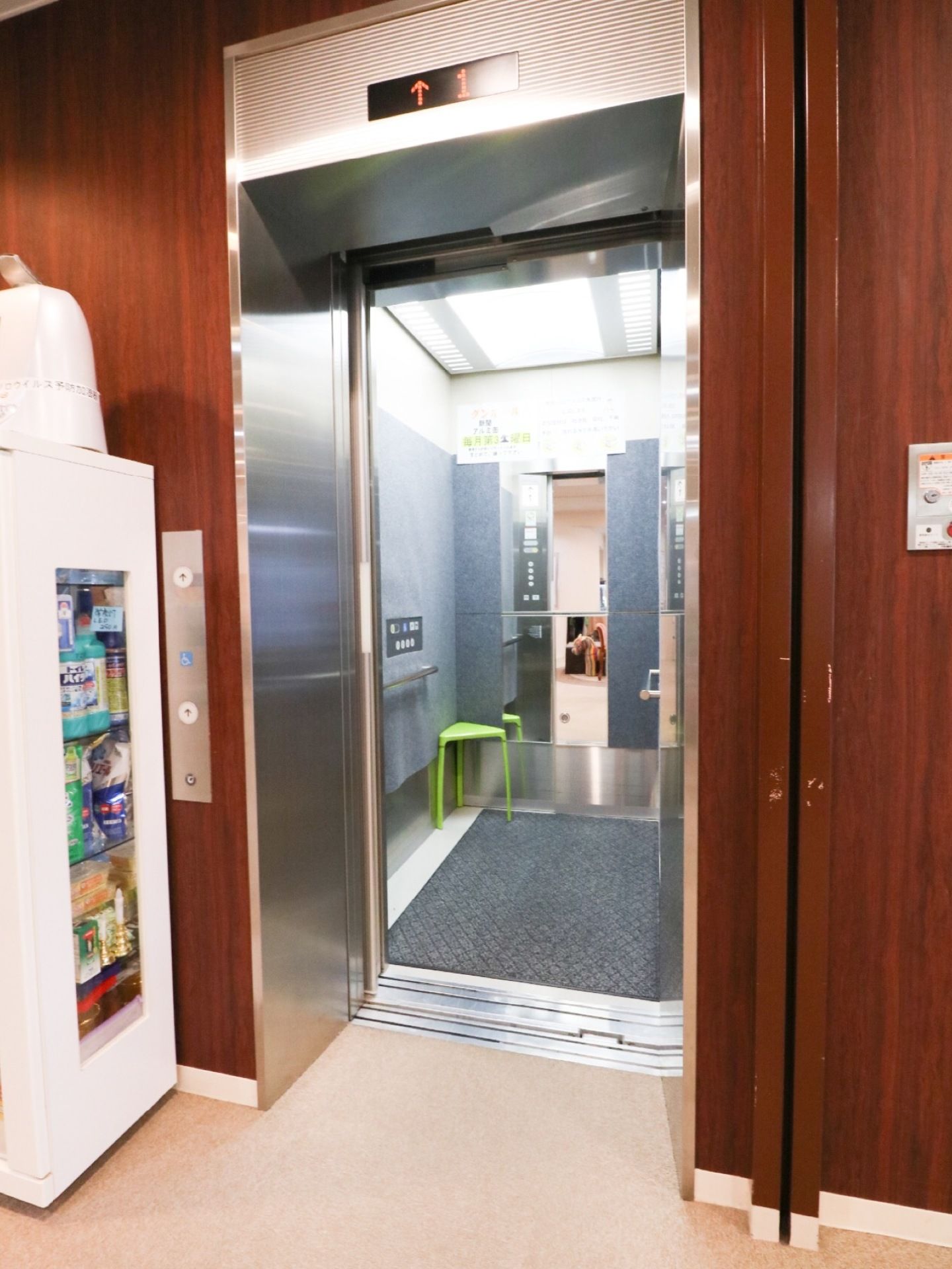 施設内に完備されているエレベーターである。１階で止まっており、ドアが開いている。床にはマットが敷かれており、奥には鏡がある。