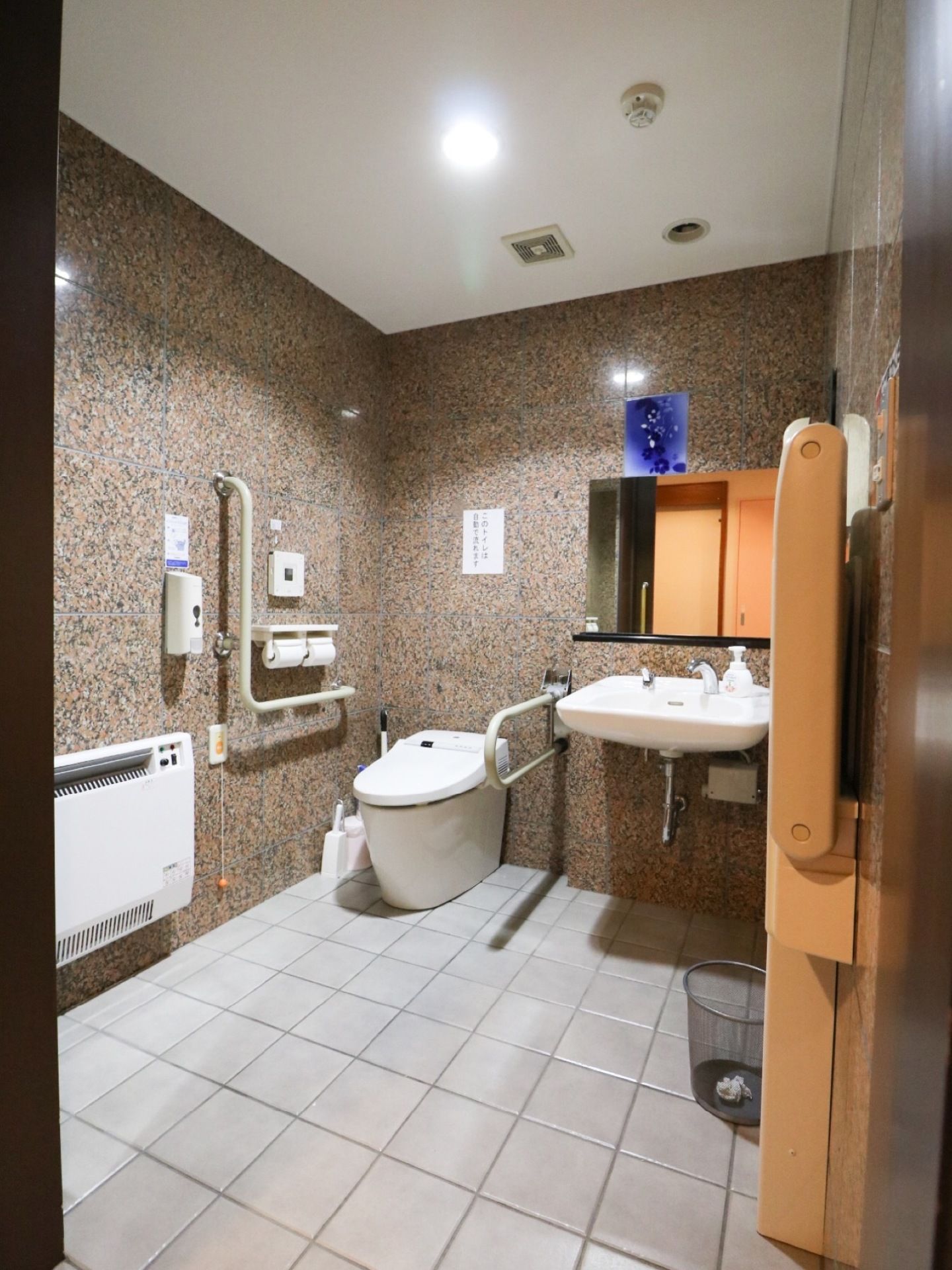 トイレはまだら模様の壁と白いタイルというデザインの、広い室内。温水洗浄便座仕様で両側に手すりがある。