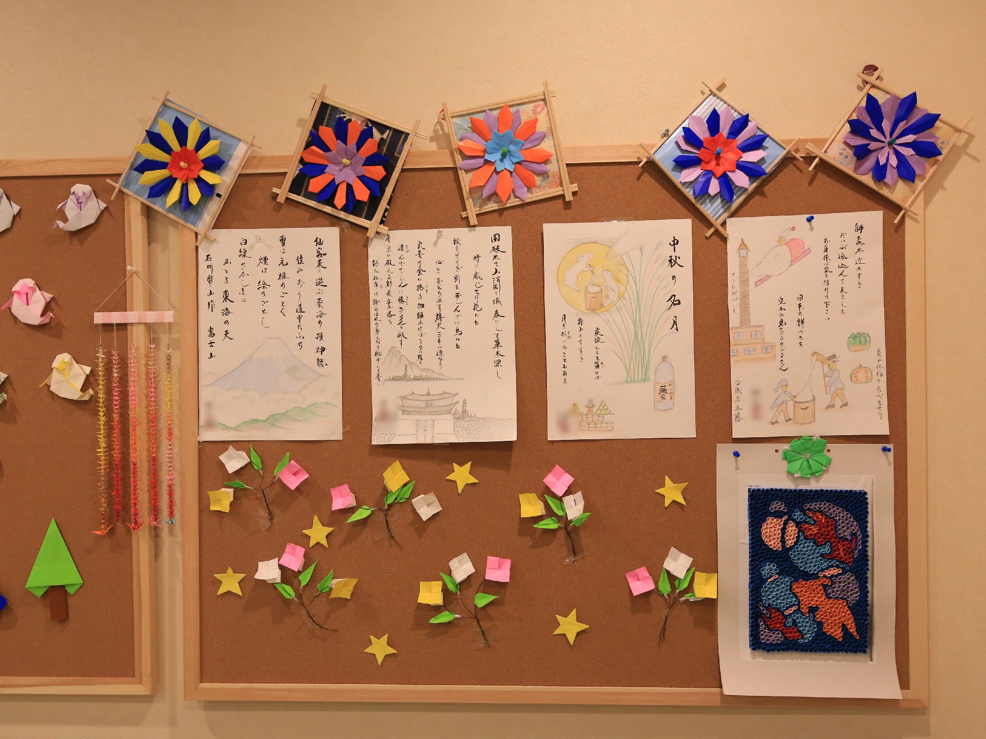 壁にコルクボードが貼ってあり、絵手紙が４枚掲示されている。空いたスペースには、折り紙で花や星が作ってあり一緒に貼られている。