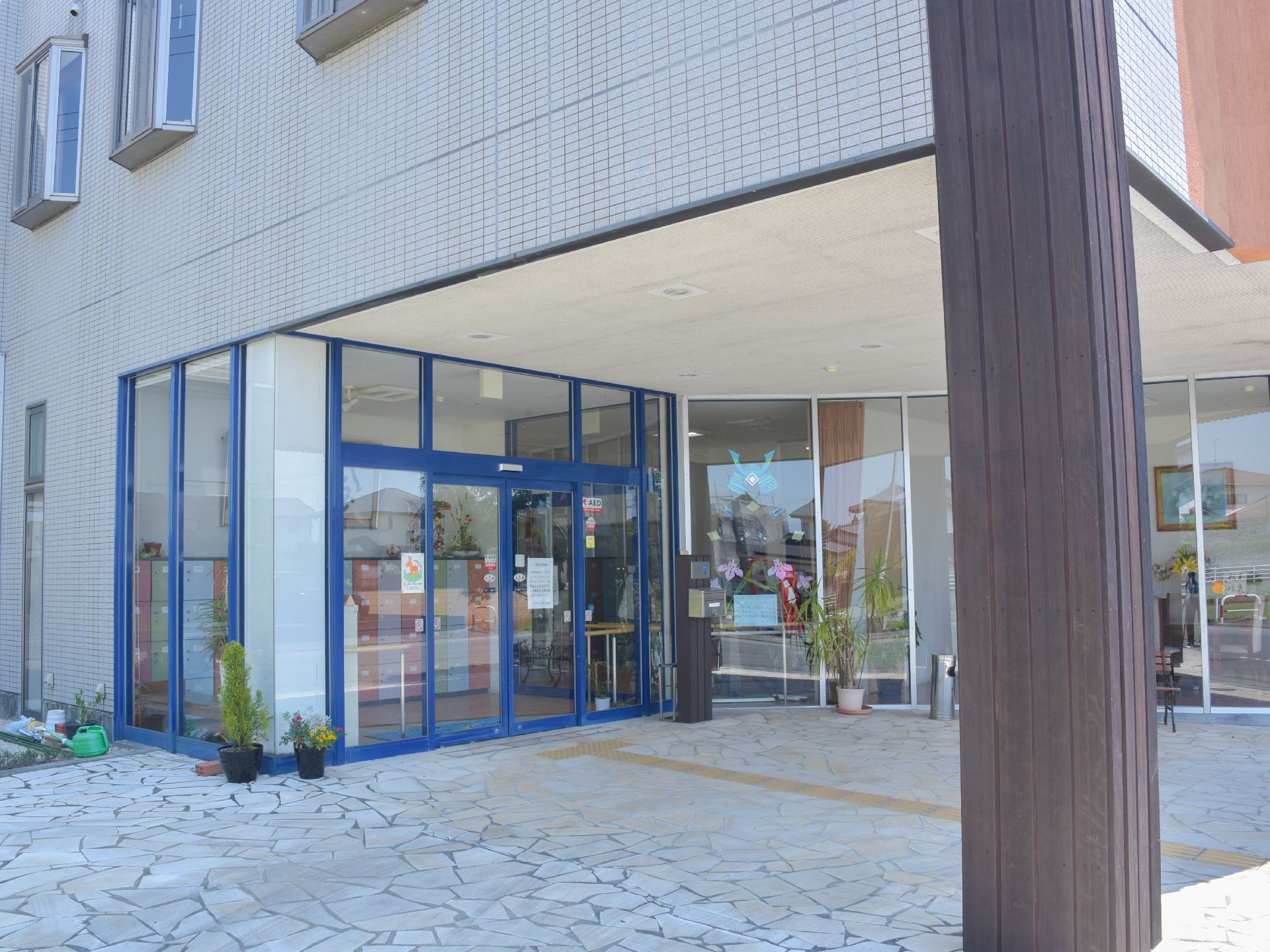 施設はグレーの外観になっていて、玄関入り口近くには点字ブロックが敷かれている。観葉植物がいくつか建物の周りに置かれている。