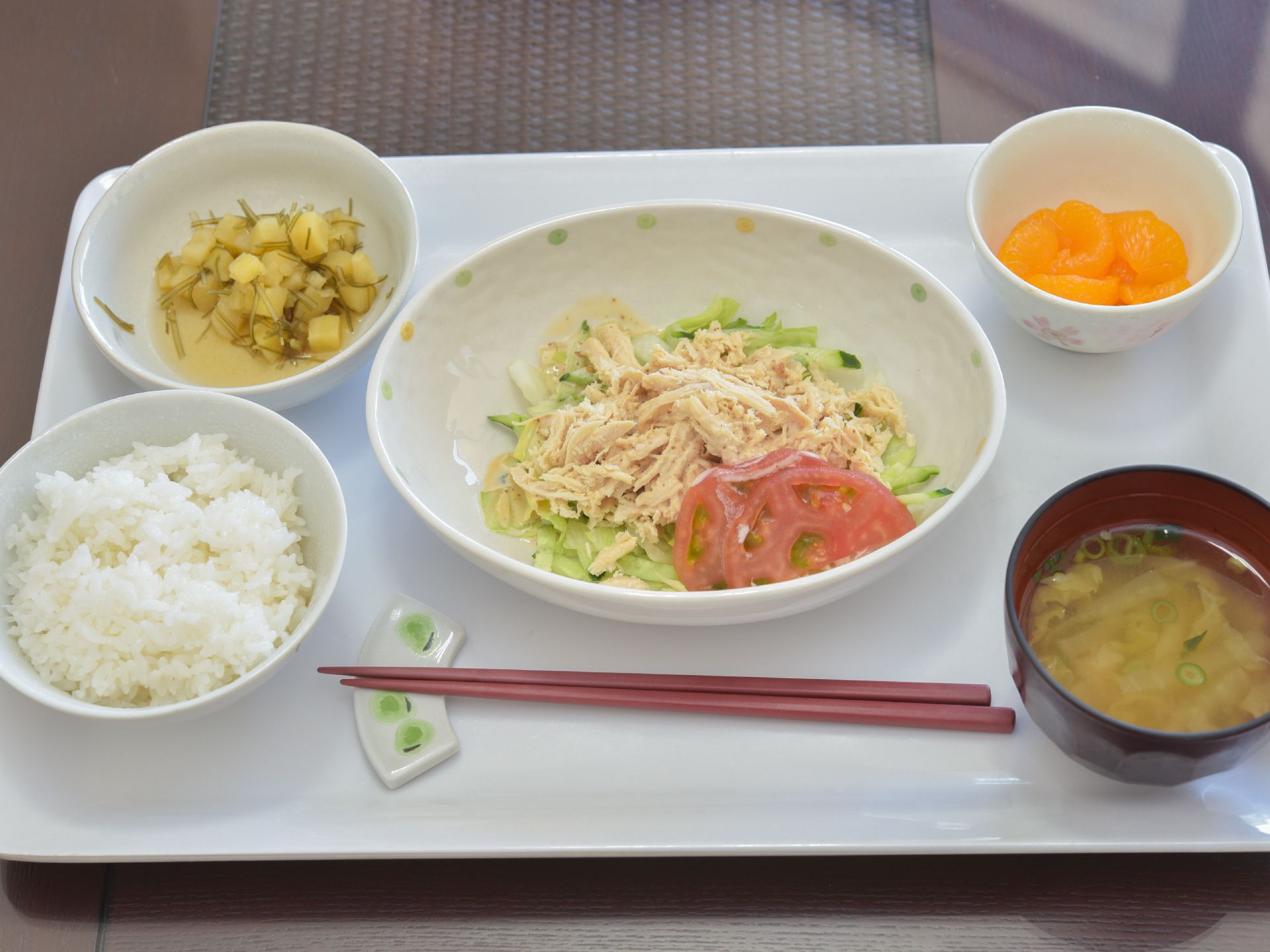 白いお盆の上には箸置きに置かれたお箸とごはんとお味噌汁、野菜を使ったおかず、サラダ、フルーツが並べられている。