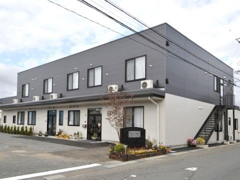 ２階建ての非常階段が設置されているグループホームである。施設前には色とりどりの花が植えられた花壇があり、看板が立てられている。