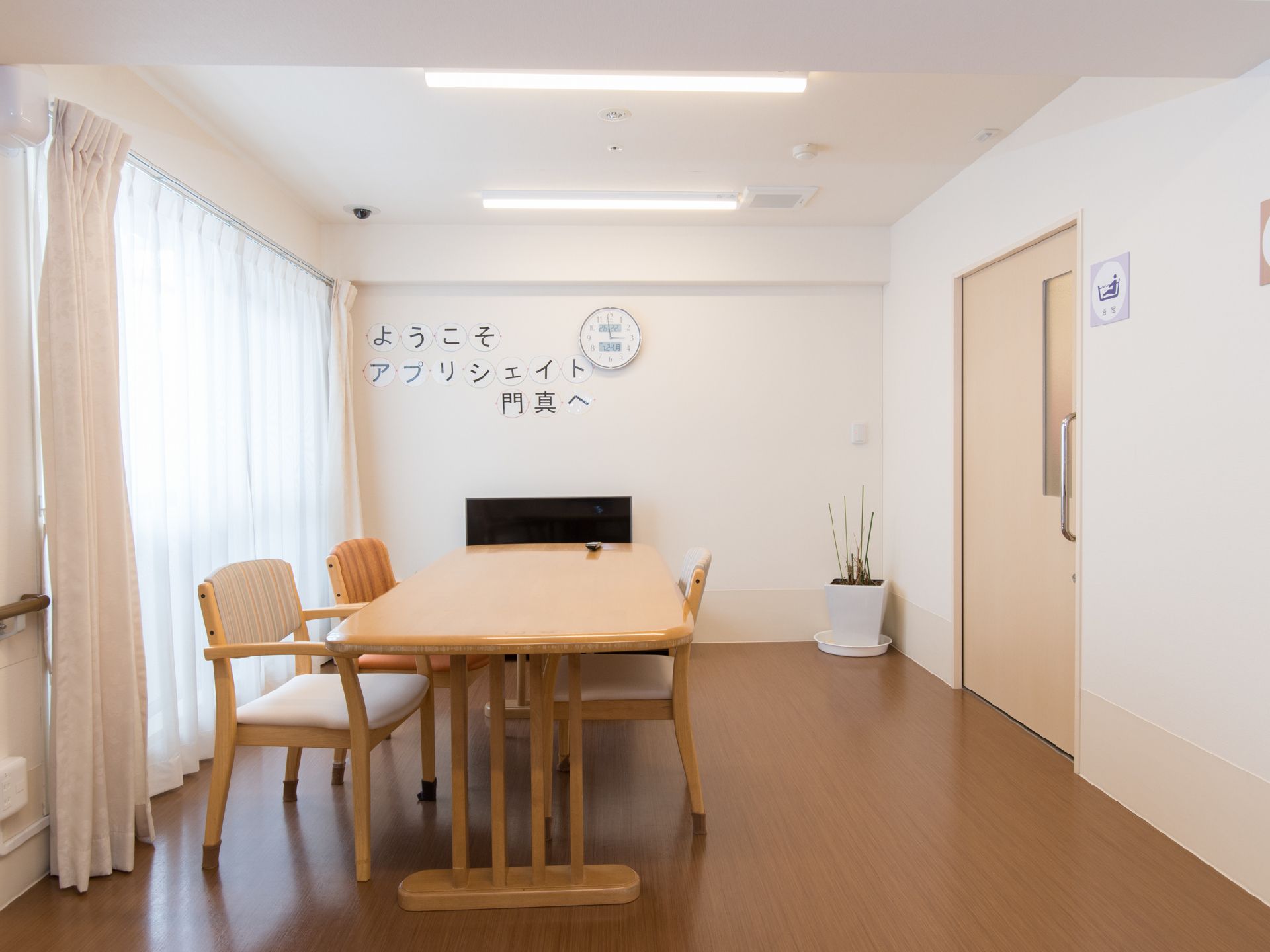 大きな窓から明るい日差しが入って来る談話室。大型テレビが設置されていて、部屋の隅には観葉植物がある。隣は浴室となっている。