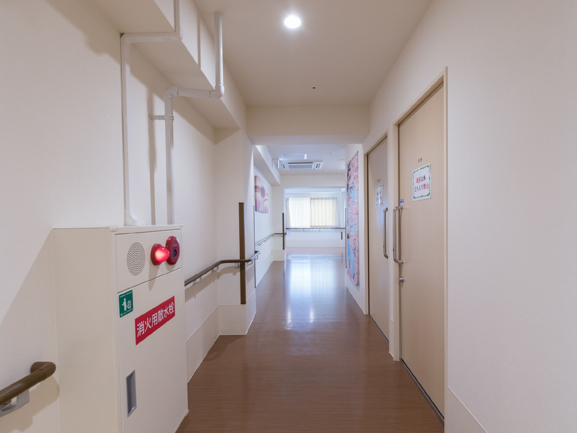 廊下には窓があり、日差しが入って明るい。非常ベルと消火用散水栓が設置されている。天井には大型のエアコンも設置されている。