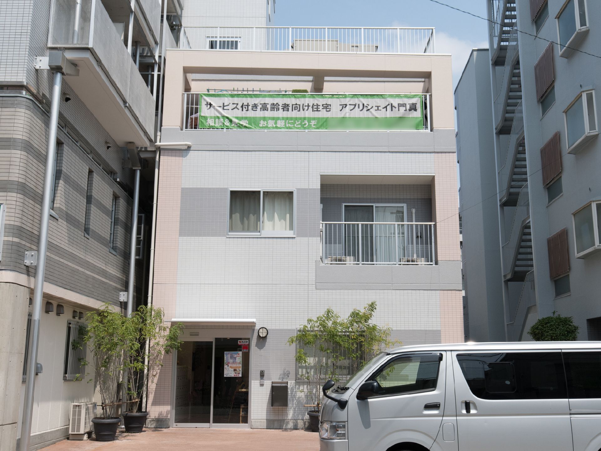 白にピンクの縁どりがされたタイル張りのビル。ところどことにバルコニーが見える。入口は1階で、手前には木の植えられた鉢植えが置かれている。