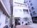 ビルに囲まれた場所に位置している。建物の前に駐車場があり車が1台止まっている。植木鉢が4つ飾られている。
