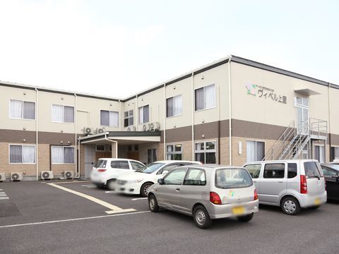 ベージュとアイボリーでまとめた２階建て建物の横には広々とした駐車場がある。建物入り口は天候に左右されない屋根付き。