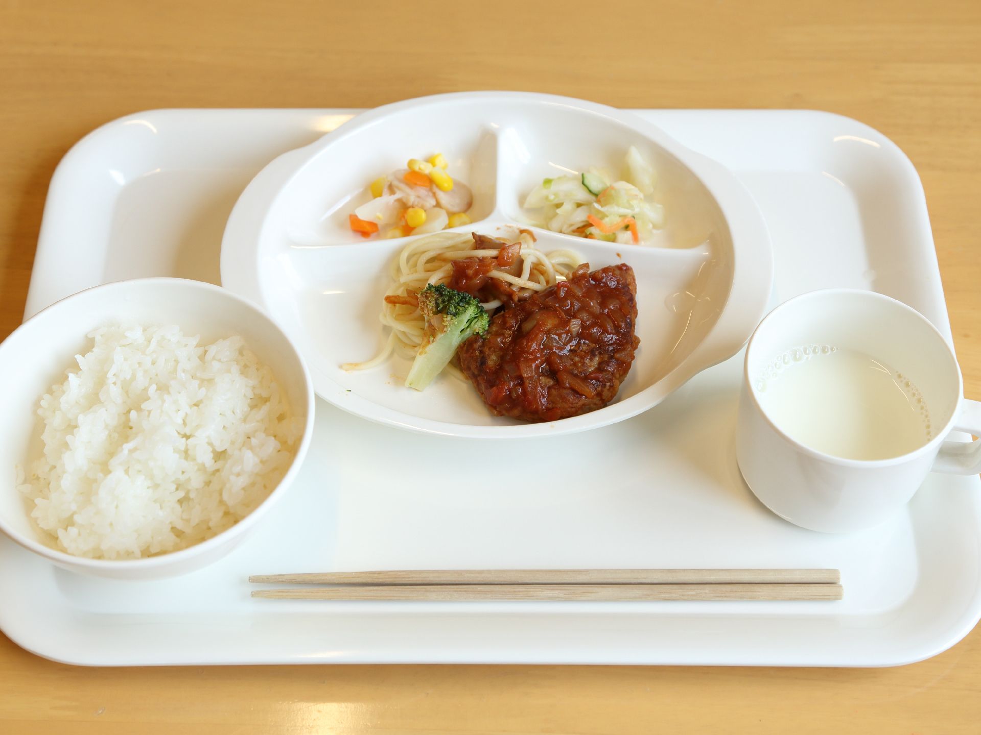 お盆の上にごはんが乗っている。仕切りがあるプレートにハンバーグ、スパゲティー、ゆでたブロッコリーなどが盛り付けてある。