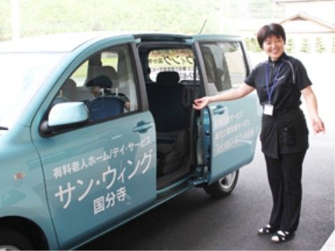 施設名称の入った送迎車の扉を開け、女性スタッフが入居者様を車内へ促すしぐさをして、にこやかに立っている。