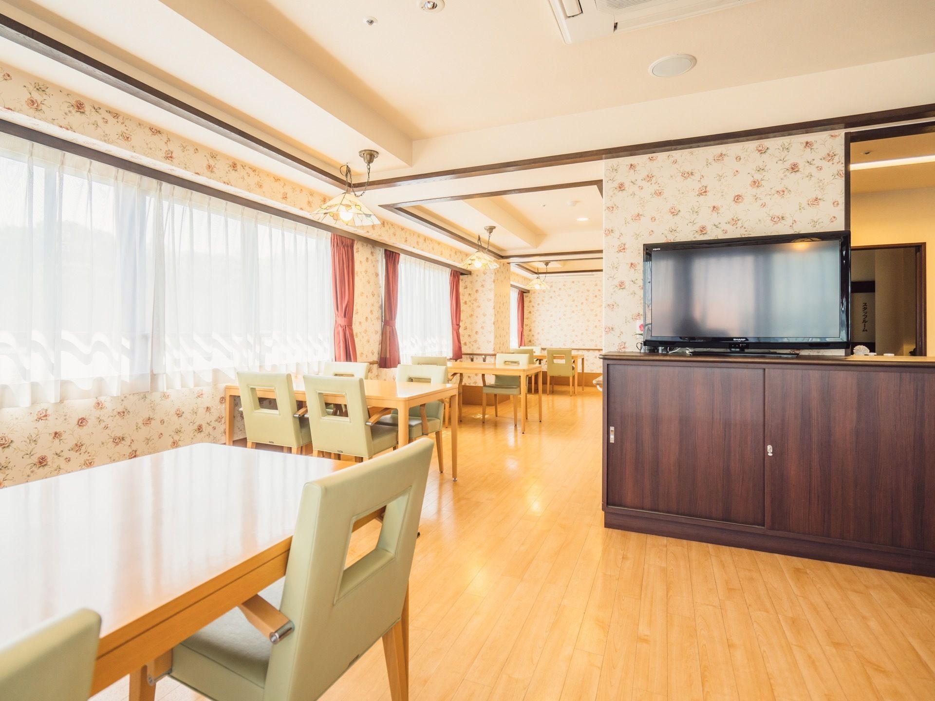 木目調の床にテレビ台、木製のテーブルと椅子、ベージュ系の花柄の壁紙と明るく家庭的な雰囲気になっている。