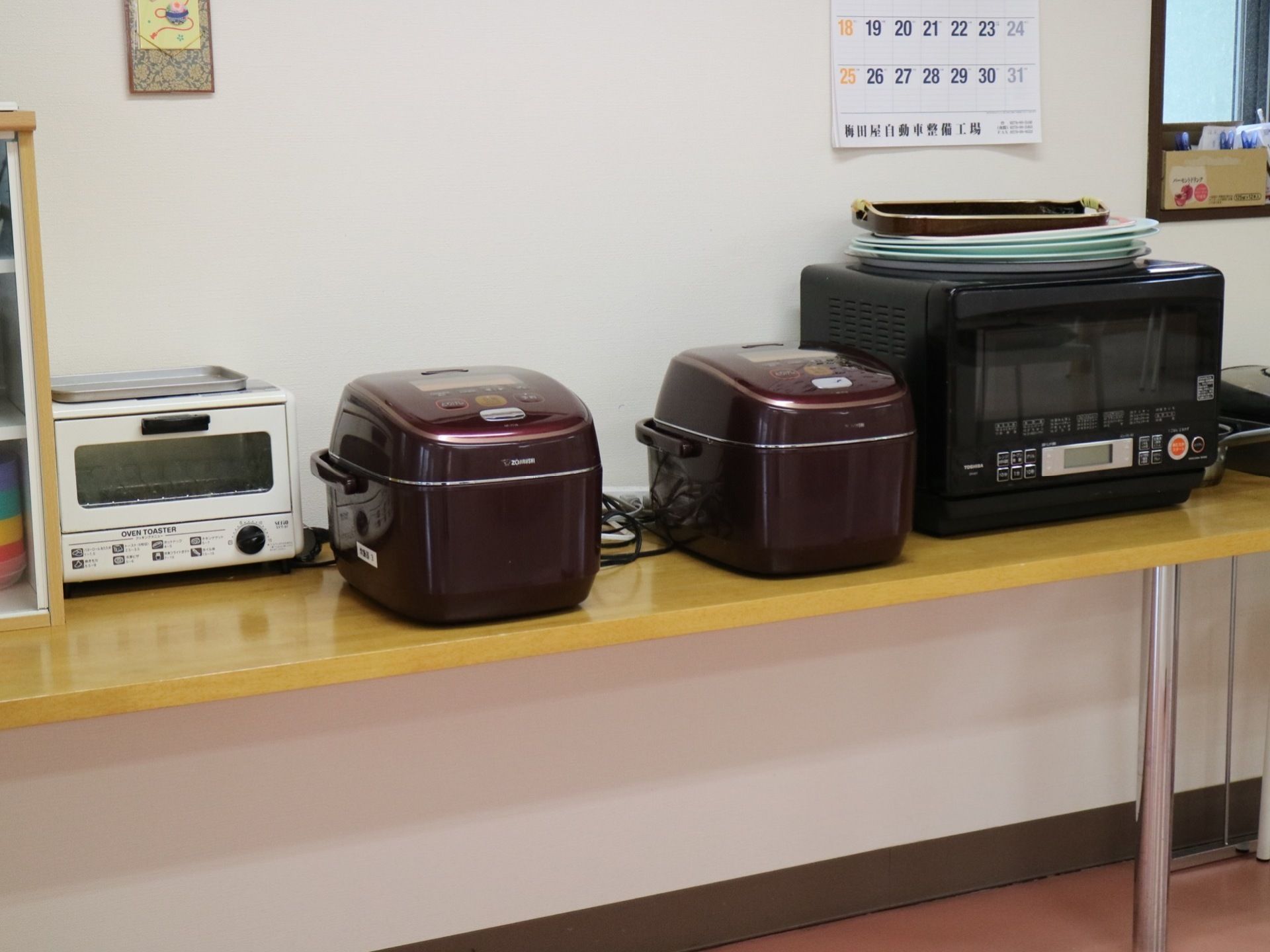 長テーブルに炊飯器や電子レンジ、トースターや食器棚が置かれている。後ろの壁には、カレンダーが掛けられている。