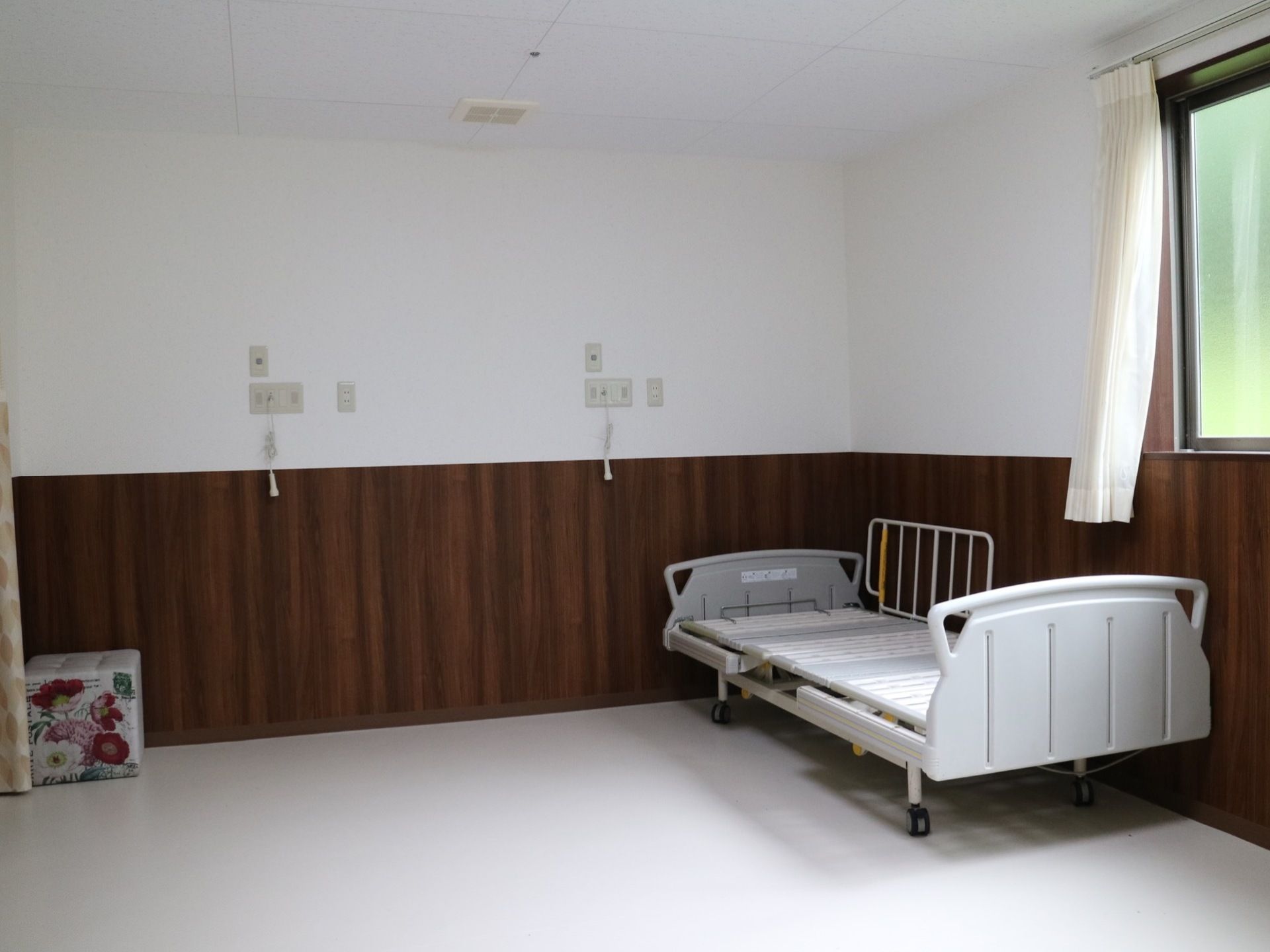 ゆとりある広々とした２人部屋である。壁にはナースコールが設置されており、窓際にはキャスター付きの介護ベッドがある。