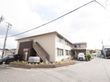 施設は2階建てで外階段がある。建物の周りには植え込みがあり、敷地内には車が停められている。部屋の窓にカーテンがかけられている。