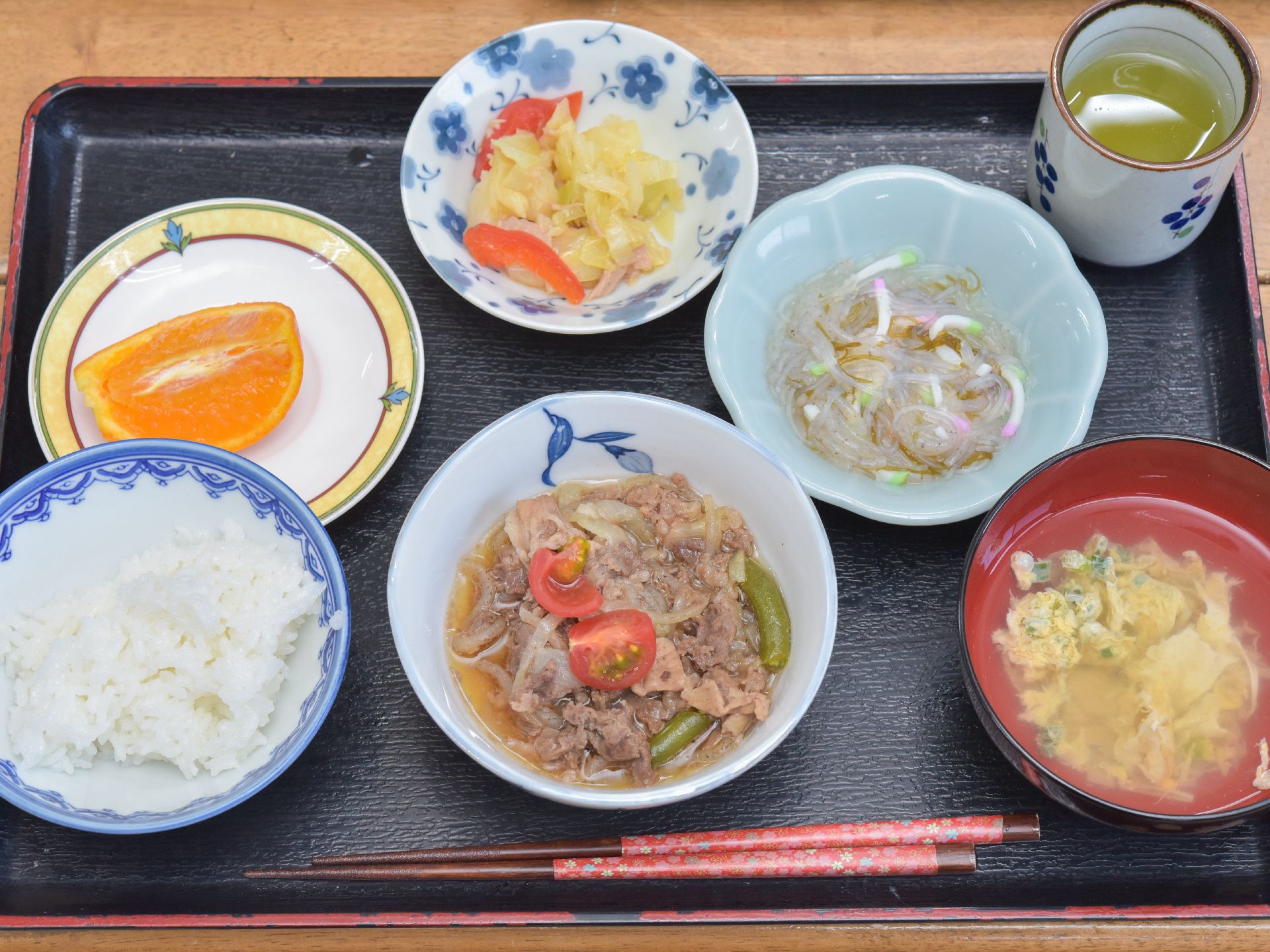 肉の煮物、野菜炒め、白滝の和え物、かき玉汁の他、デザートのオレンジ等、栄養のバランスを考えた献立が配膳されている。