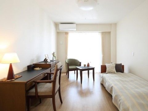 開放的な居室。エアコンで取り付けられている。机とチェストがあり、ランプや小物が設置されている。ベッドの上にクッションが置かれている。