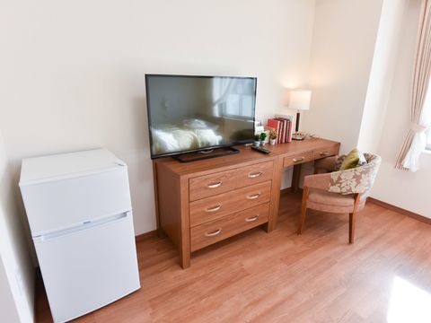 引き出しの多い机の上に、テレビと照明が設置されている。椅子に腰かけて、机で読書や書き物をすることができる。左側には冷蔵庫が設置されている。