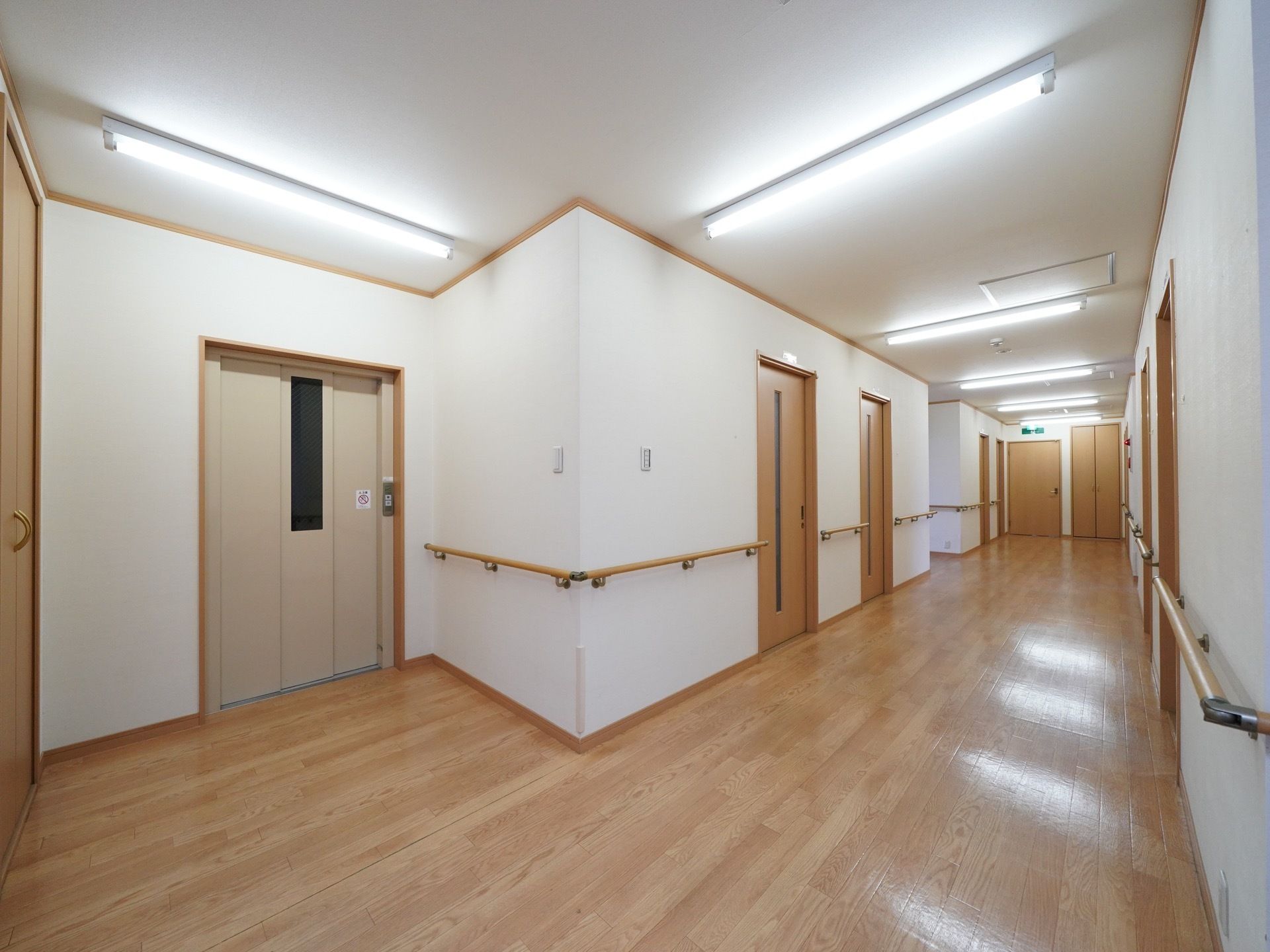 両サイドに入居者様の居室が並んでいる通路がある。壁には手すりが設置されており、エレベーターホールへと繋がっている。