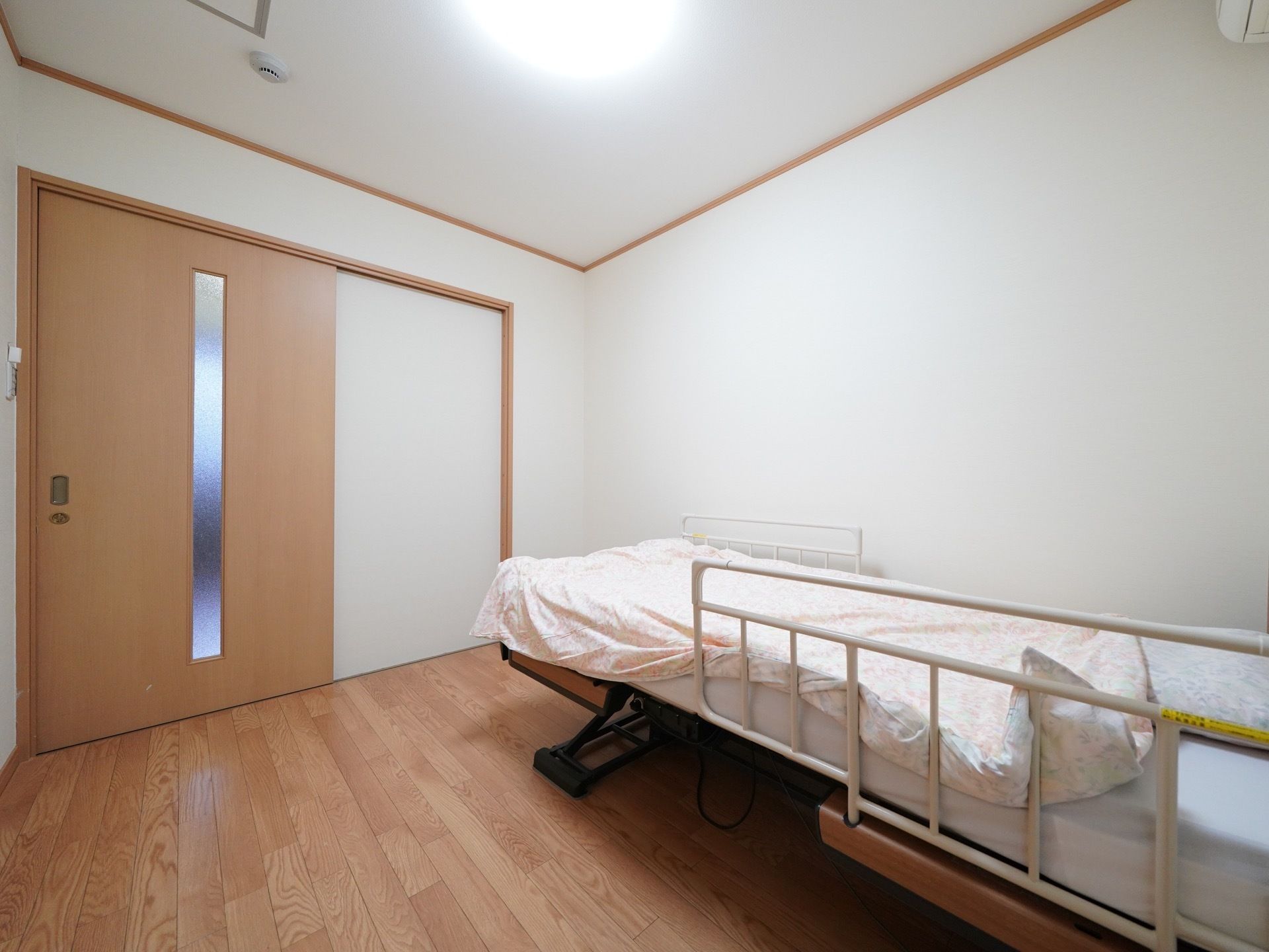 すりガラスのついたベージュの引き戸のある入居者様の居室。木のぬくもりを感じさせる材質の床と、壁際には介護用ベッドが置かれている。