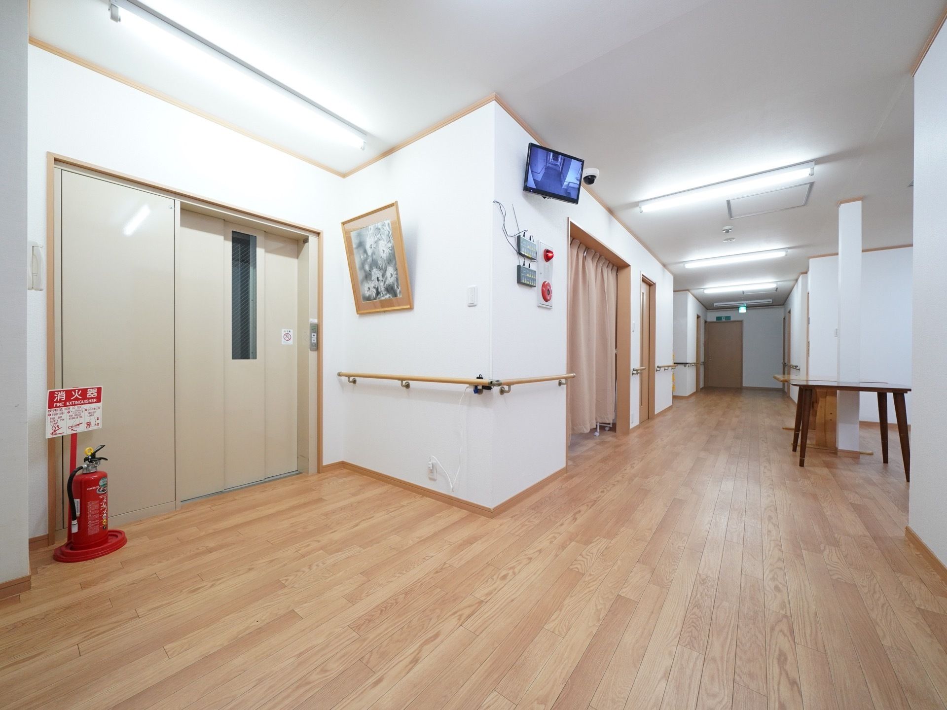 居室と談話室のある一角にエレベーターホールがある。壁には額縁が飾られており、施設内を映すモニターが天井近くに設置されている。