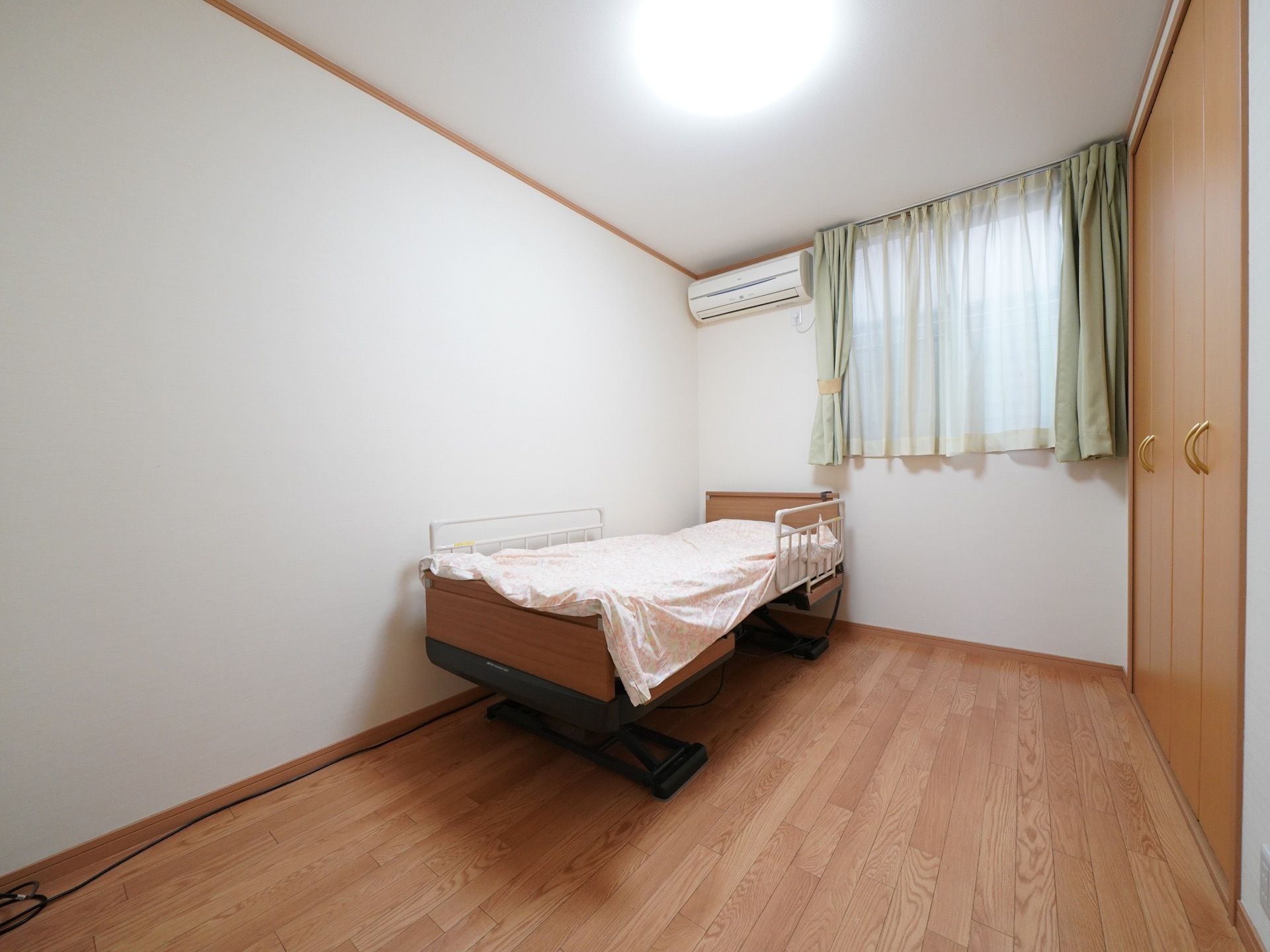 カーテンの掛かっている窓がある入居者様の居室。壁には大きなクローゼットの扉がある。介護用ベッドが置かれている。