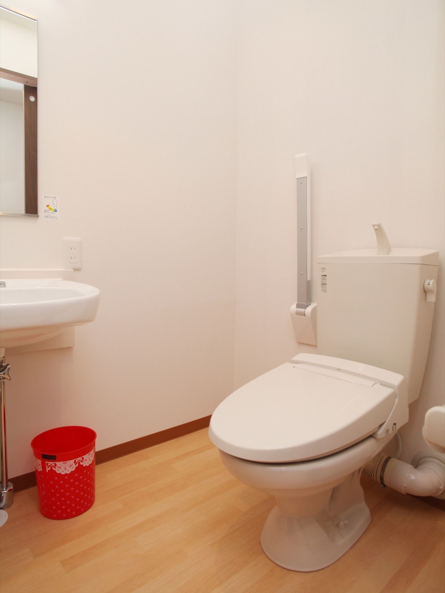 トイレは白い内装できれいな印象がある。洗面台の横の壁にはコンセントが付いており、下には赤いゴミ箱がある。