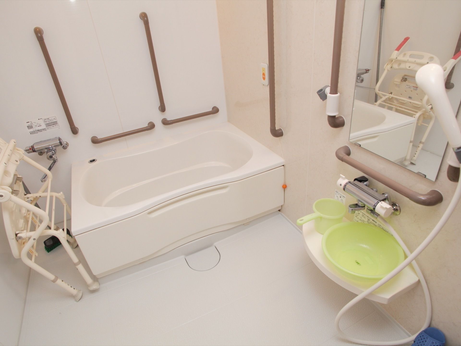 白いユニットバスで、壁には茶色の手すりが設置されている。洗面器や手桶、折り畳みのシャワーチェアが備えてある。
