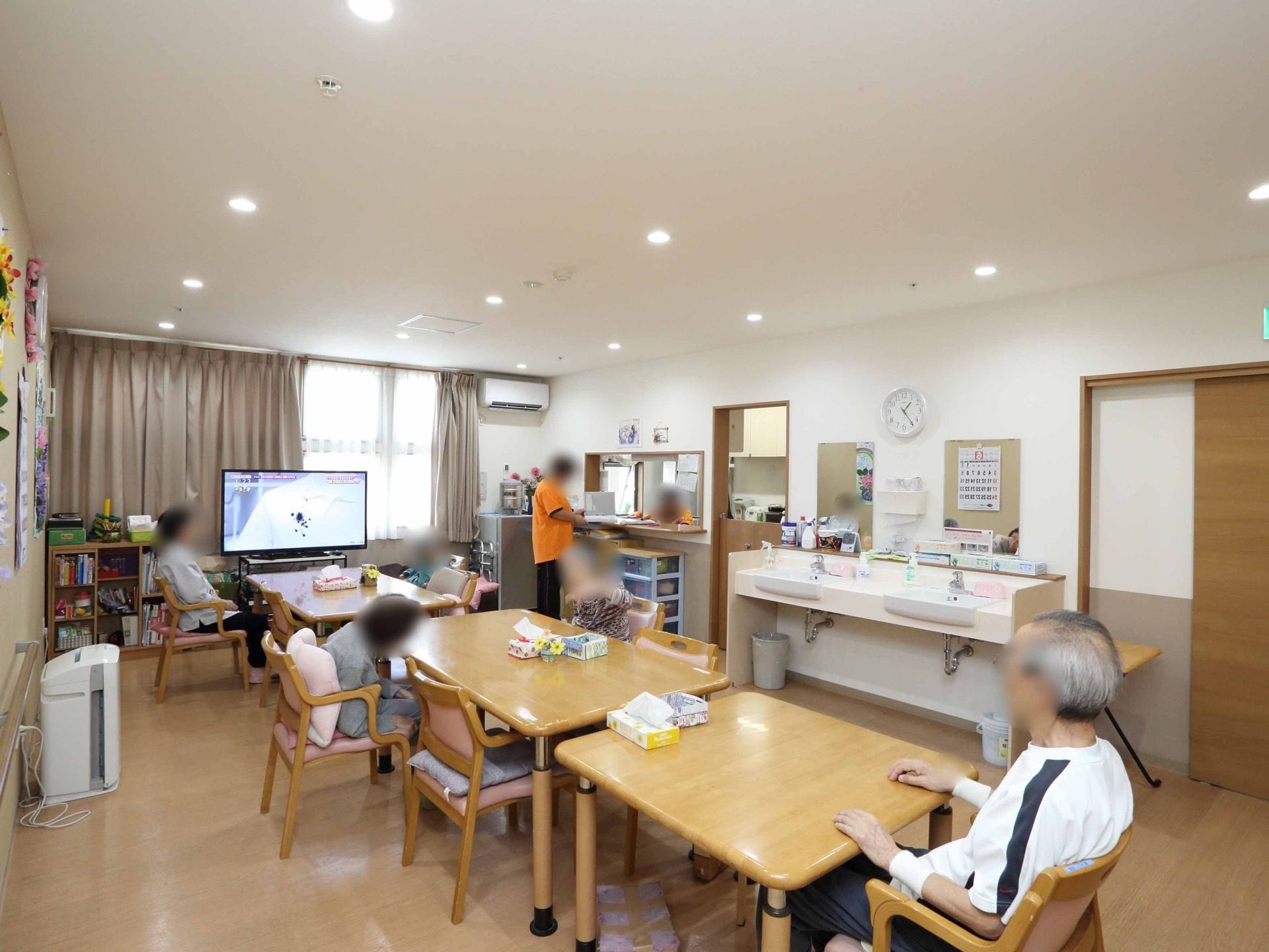 リビング横にカウンターキッチンがある。窓際にテレビや本棚が置かれており、入居者様が椅子に座って思い思いの時間を過ごしている。