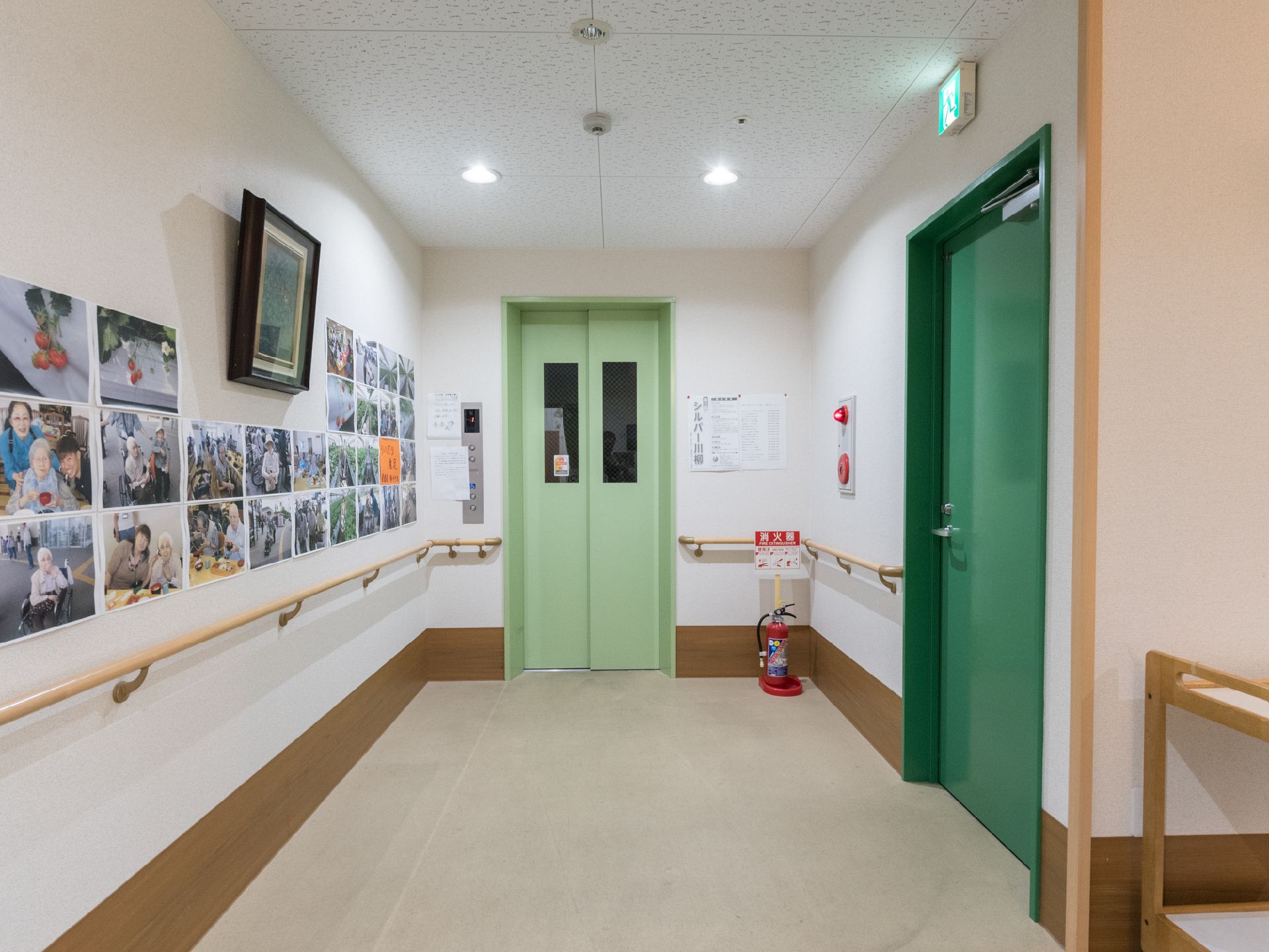 入居者様達が写った写真がたくさん貼られた手すり付きの壁がある。近くには黄緑色のエレベーターが1基設置されている。