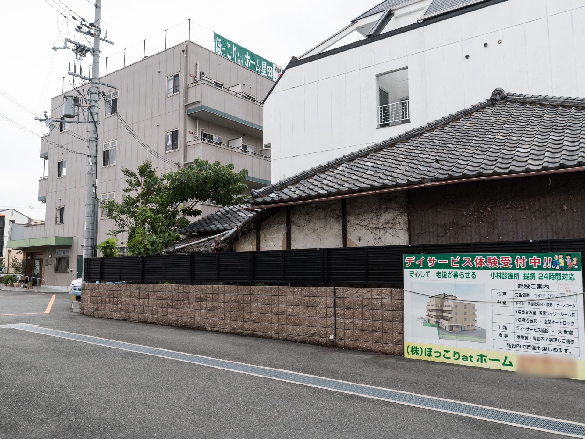 白い建物の前に瓦屋根の建物と塀があり、施設の大きな看板が取り付けられている。近くの白い建物の屋上にも施設の看板がある。