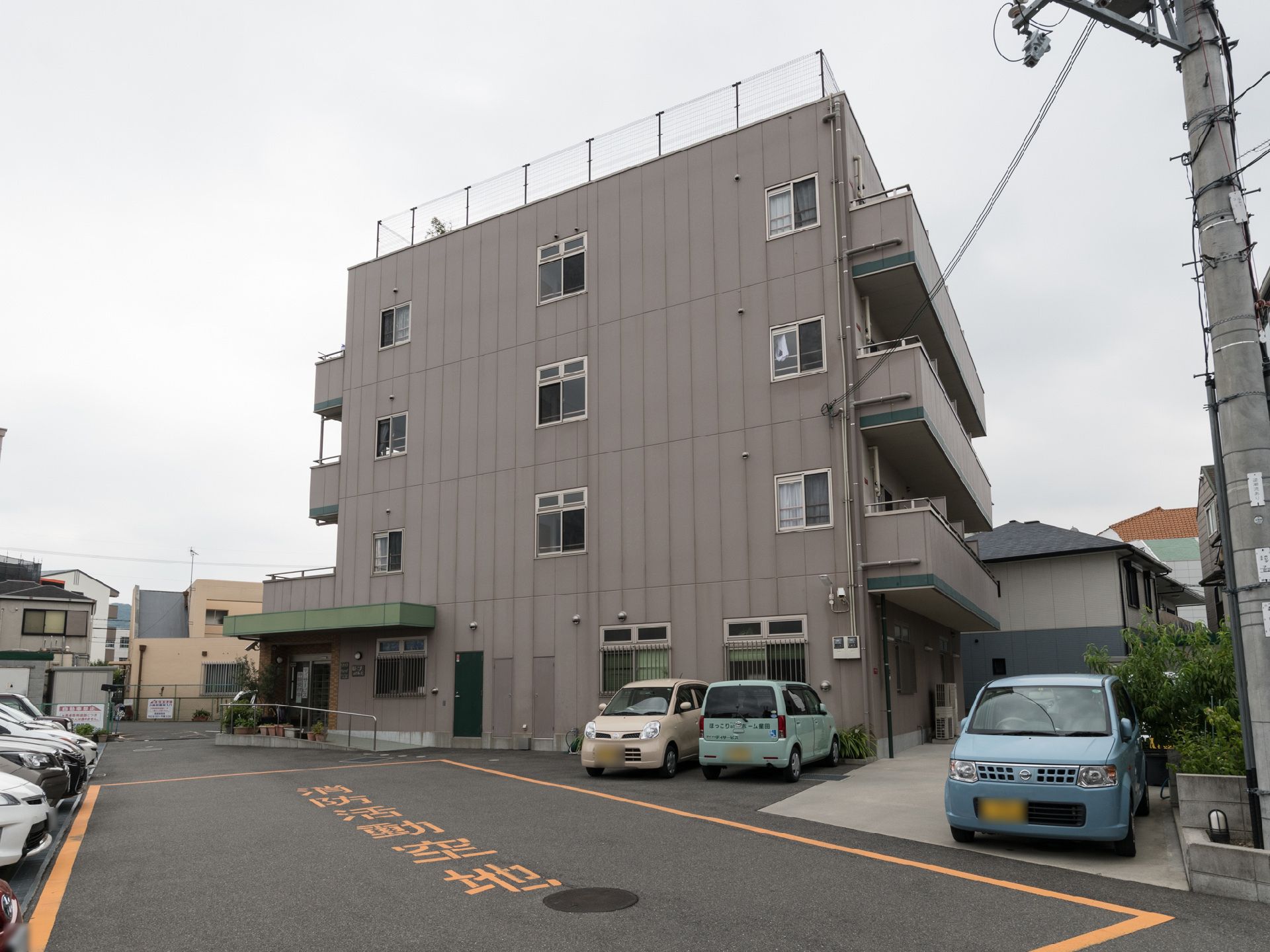 茶色い4階建ての施設付近には車の駐車スペースがある。そこには何台かの車が停まっており、真ん中には消防活動空地が設置されている。