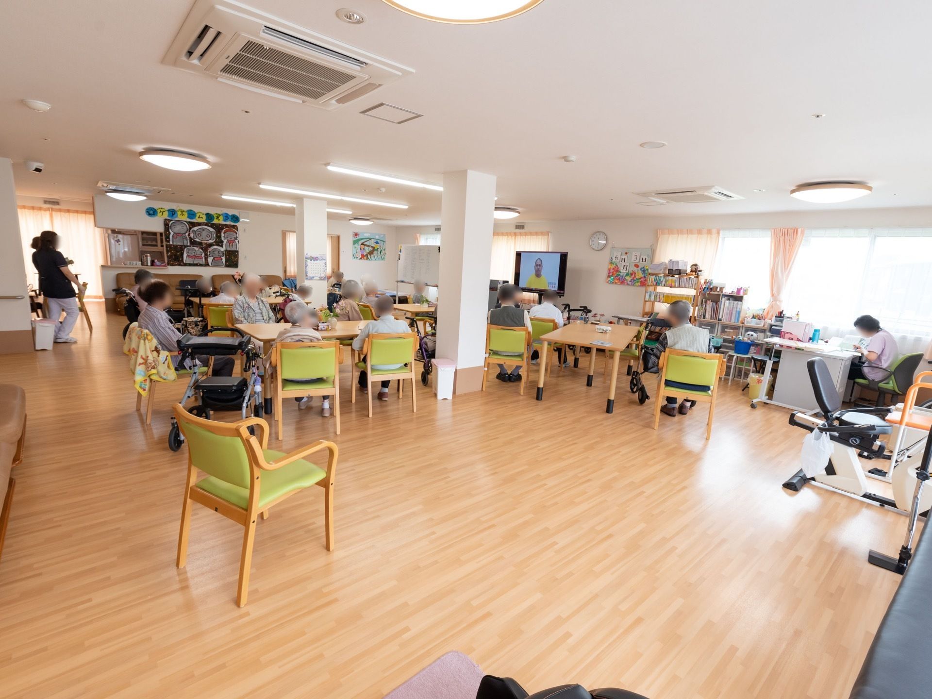 たくさんの入居者様が、椅子に座ってテレビを見ている。右端には事務用デスクがあり、女性スタッフが作業している。