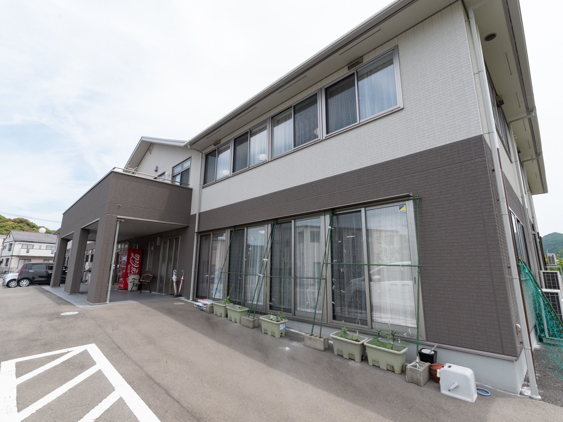 施設の外壁デザインは、グレー系の落ち着いた印象である。玄関右側の通路には、数多くのプランターが並べて置いてある。