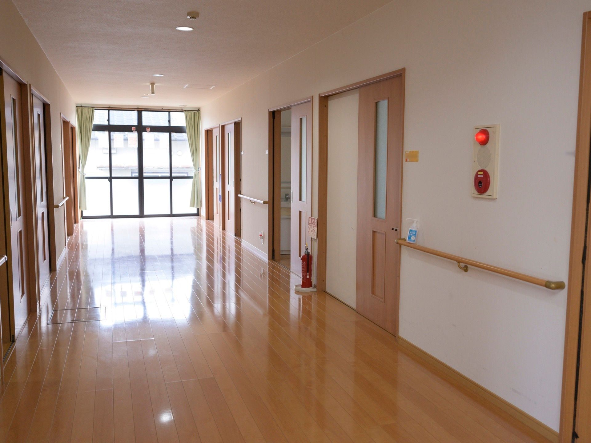突き当たりに掃き出し窓のある明るい廊下が続いている。両側には居室が並んでおり、壁には手すりが付いている。