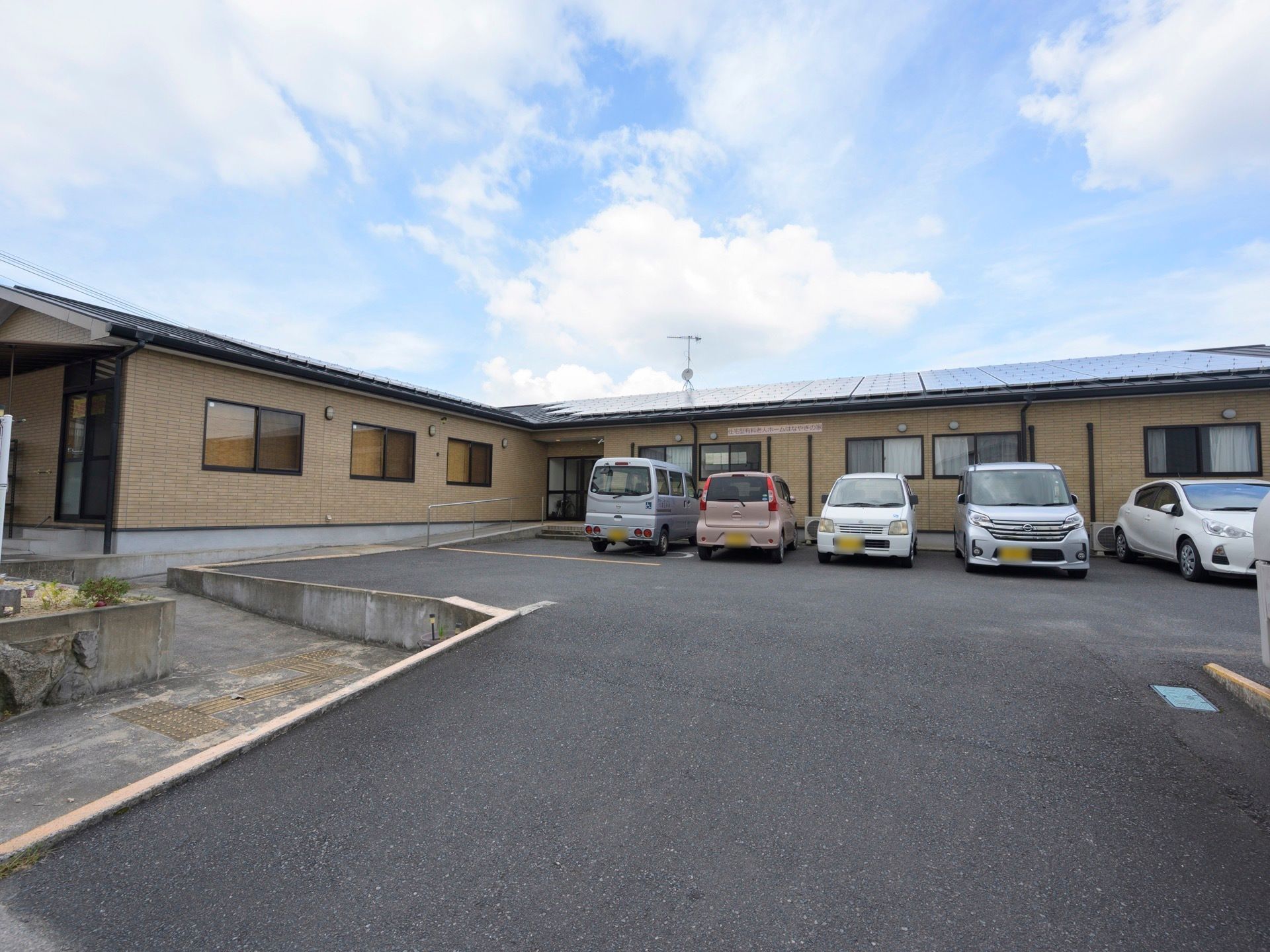 平屋建ての住宅があり、手前はアスファルトで舗装された駐車スペースになっている。車が５台、並んで停められている。