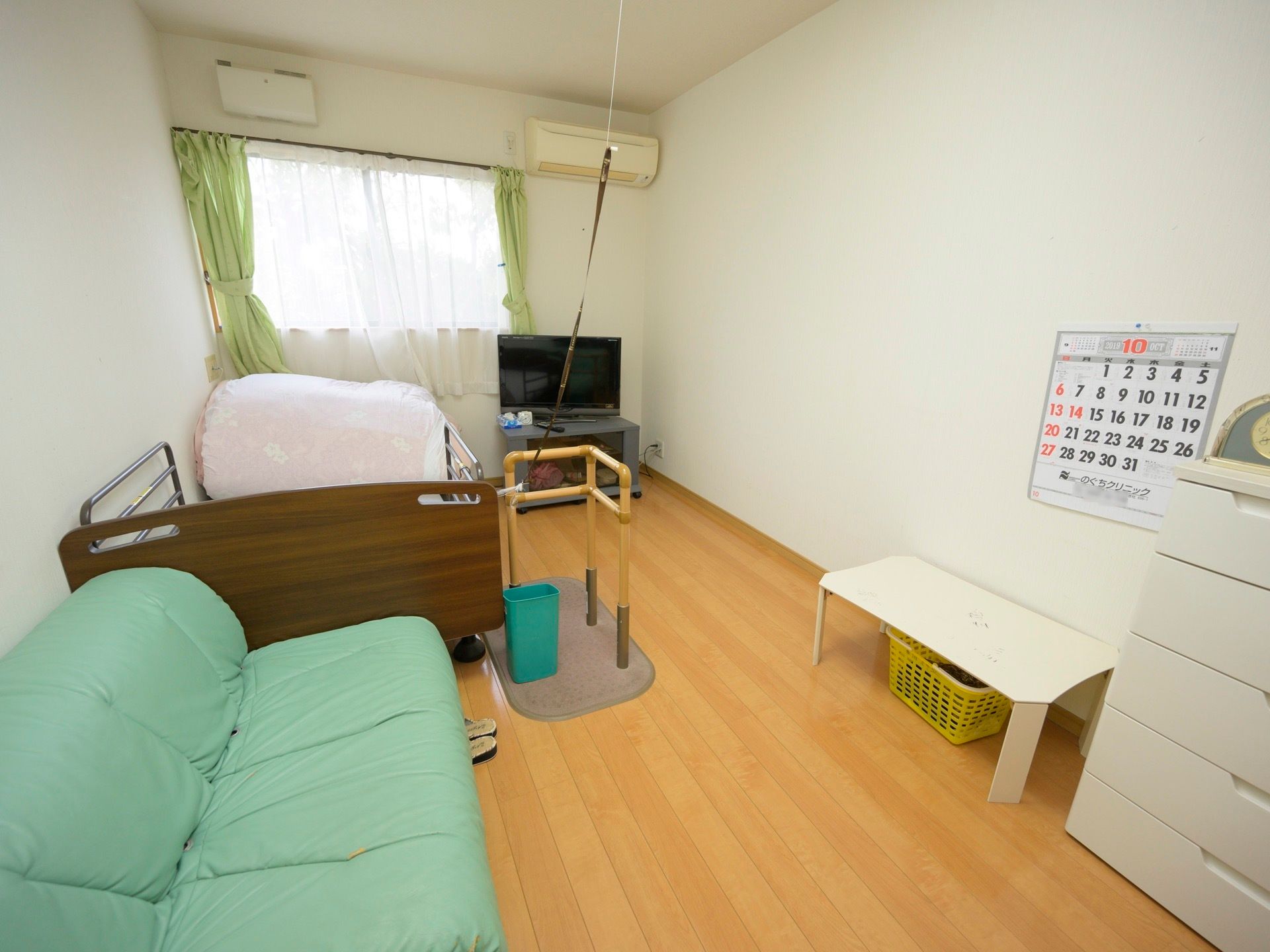 フローリングの居室に、薄い緑のソファと電動ベッド、テレビ、ミニテーブル、チェストなどが置かれている。