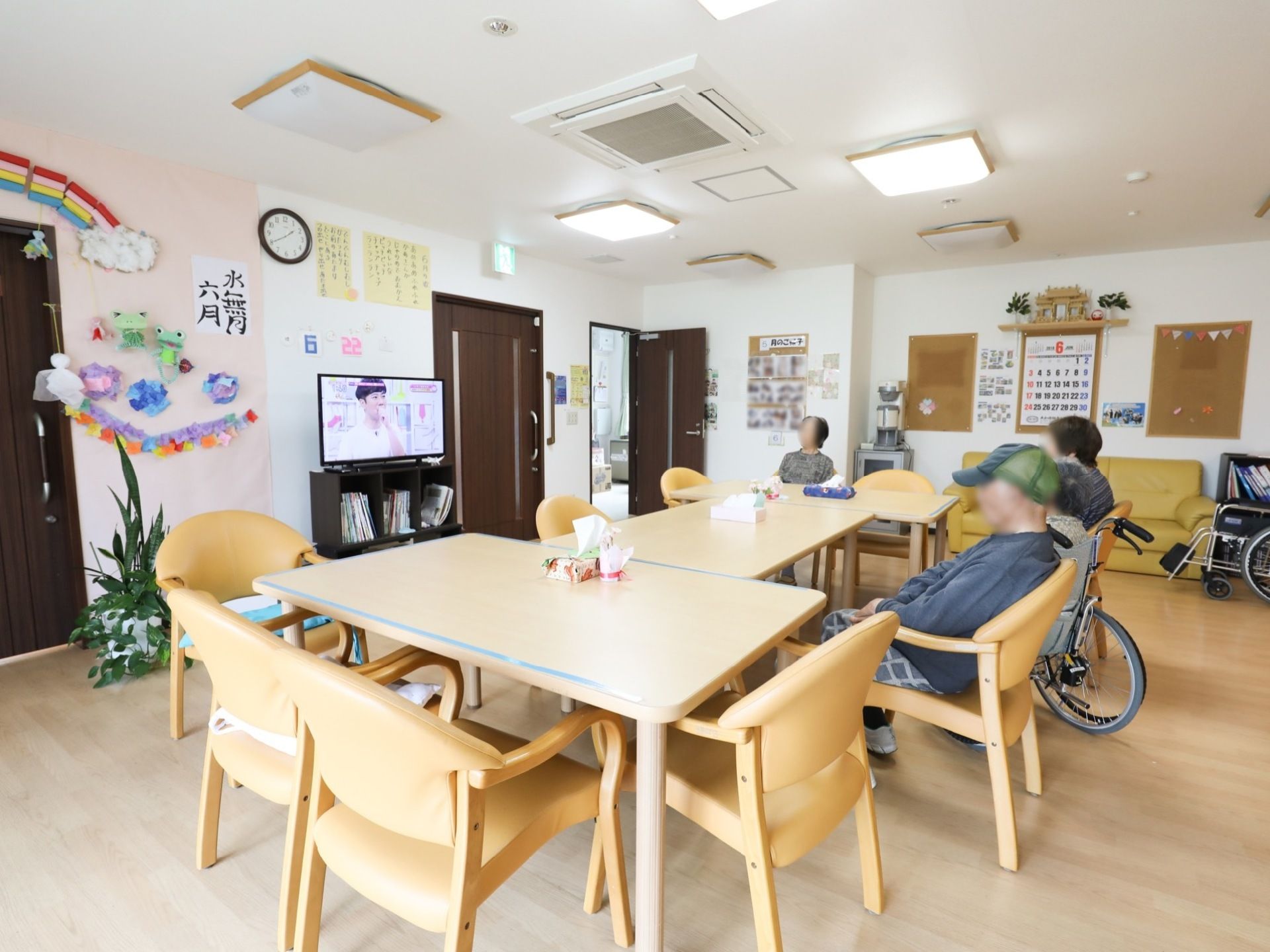 ダイニングテーブルが３台並べられている。入居者様が三名テレビを見ている。さまざまな掲示物が壁に貼られている。