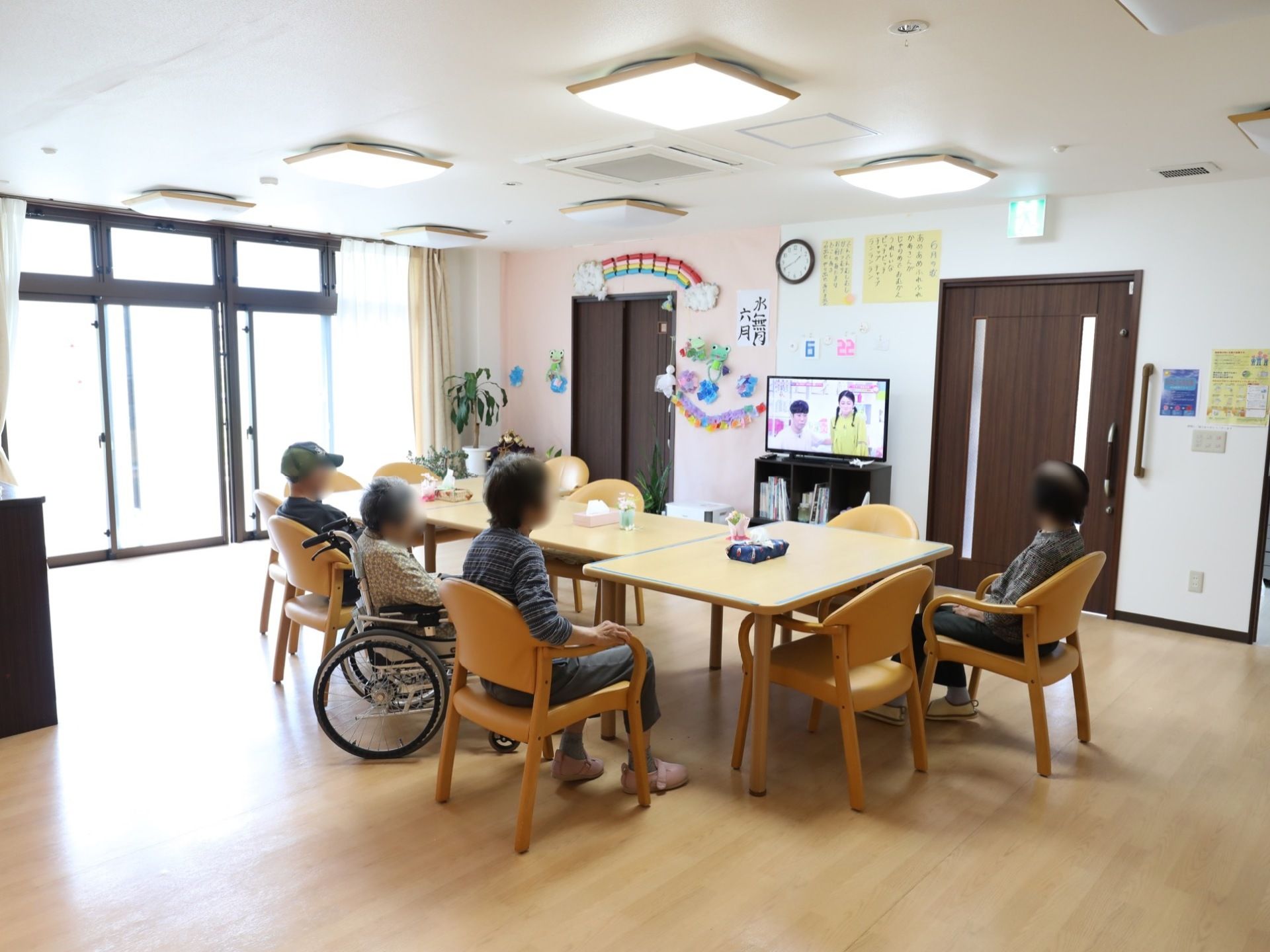 共有スペースでは、女性の車椅子をご利用の方と数名の入居者様が、設置されているテーブルセットの椅子に腰掛け、一緒にテレビをご覧になっている。