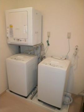 洗濯機が二台置かれ、向かって左側の洗濯機の上には乾燥機も設置されている。床はフローリング、壁は白色である。