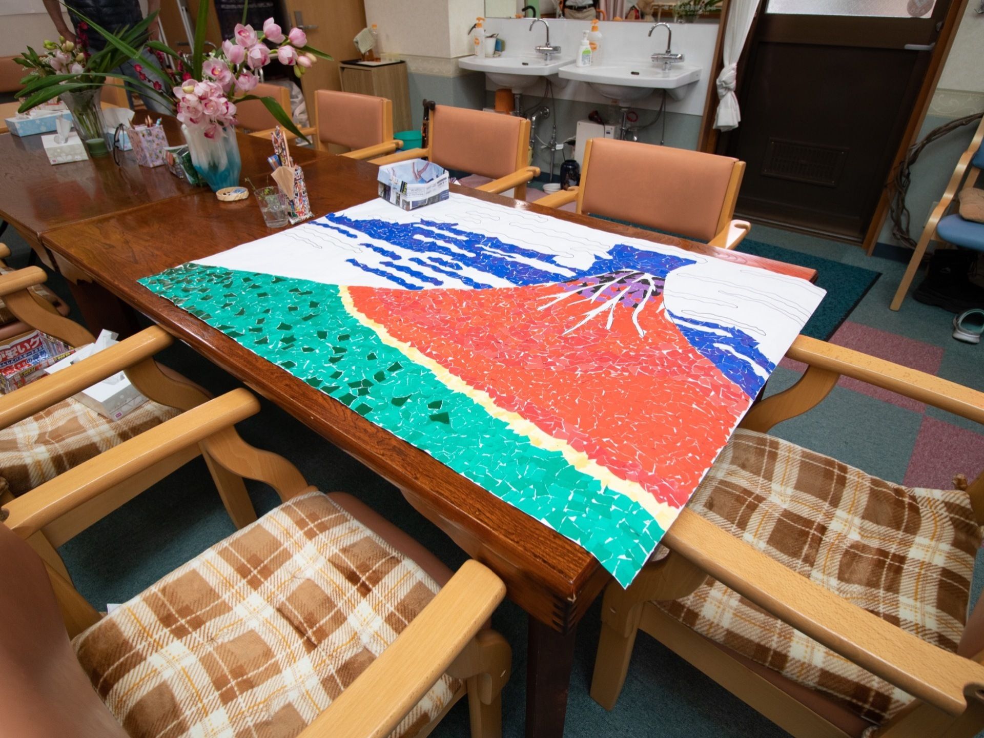 アットホームな雰囲気の共有スペースである。テーブルの上には、制作中のちぎり絵の作品が置かれている。洗面台が２台設置されている。