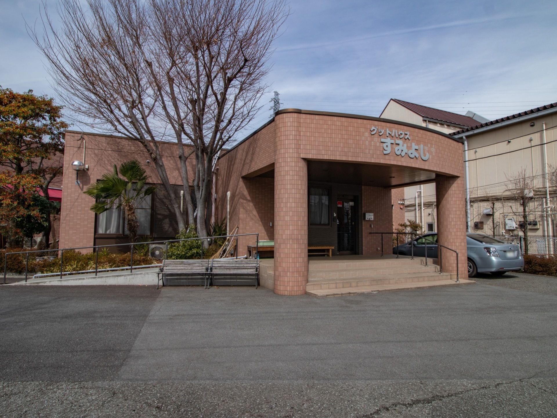 茶色の平屋の建物で、玄関の上には広く屋根が設置されている。建物周辺には、樹木がたくさん植えられており、自然を感じていただける。