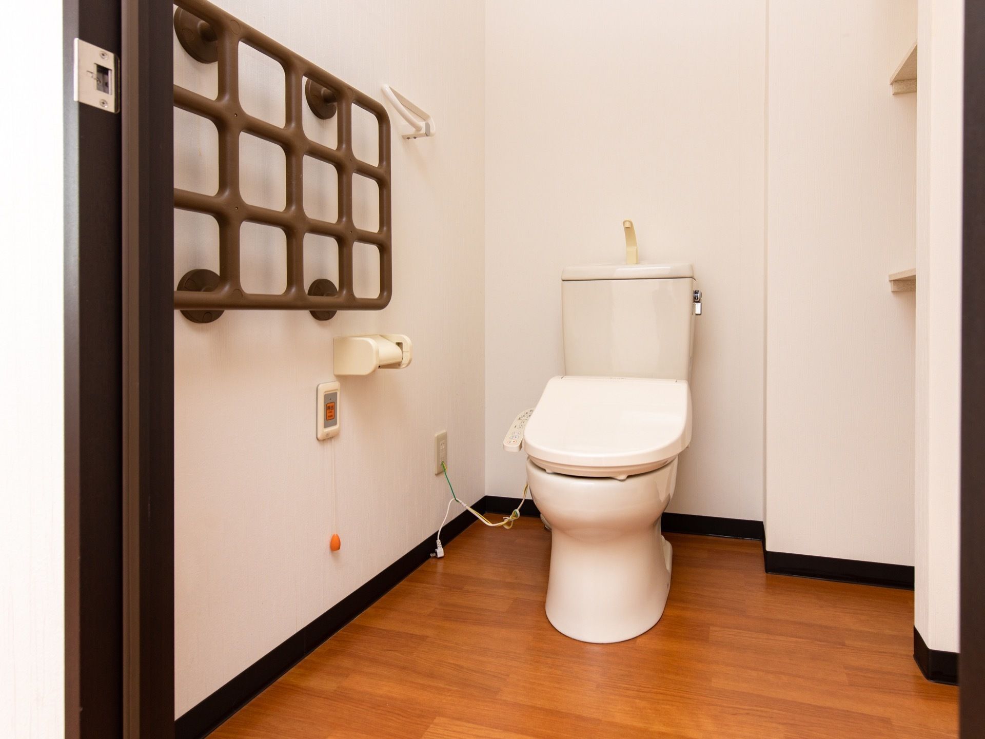 横幅が広いトイレスペースに洋式便器が据えられている。左側の壁面に、格子状のグリップやナースコールがある。