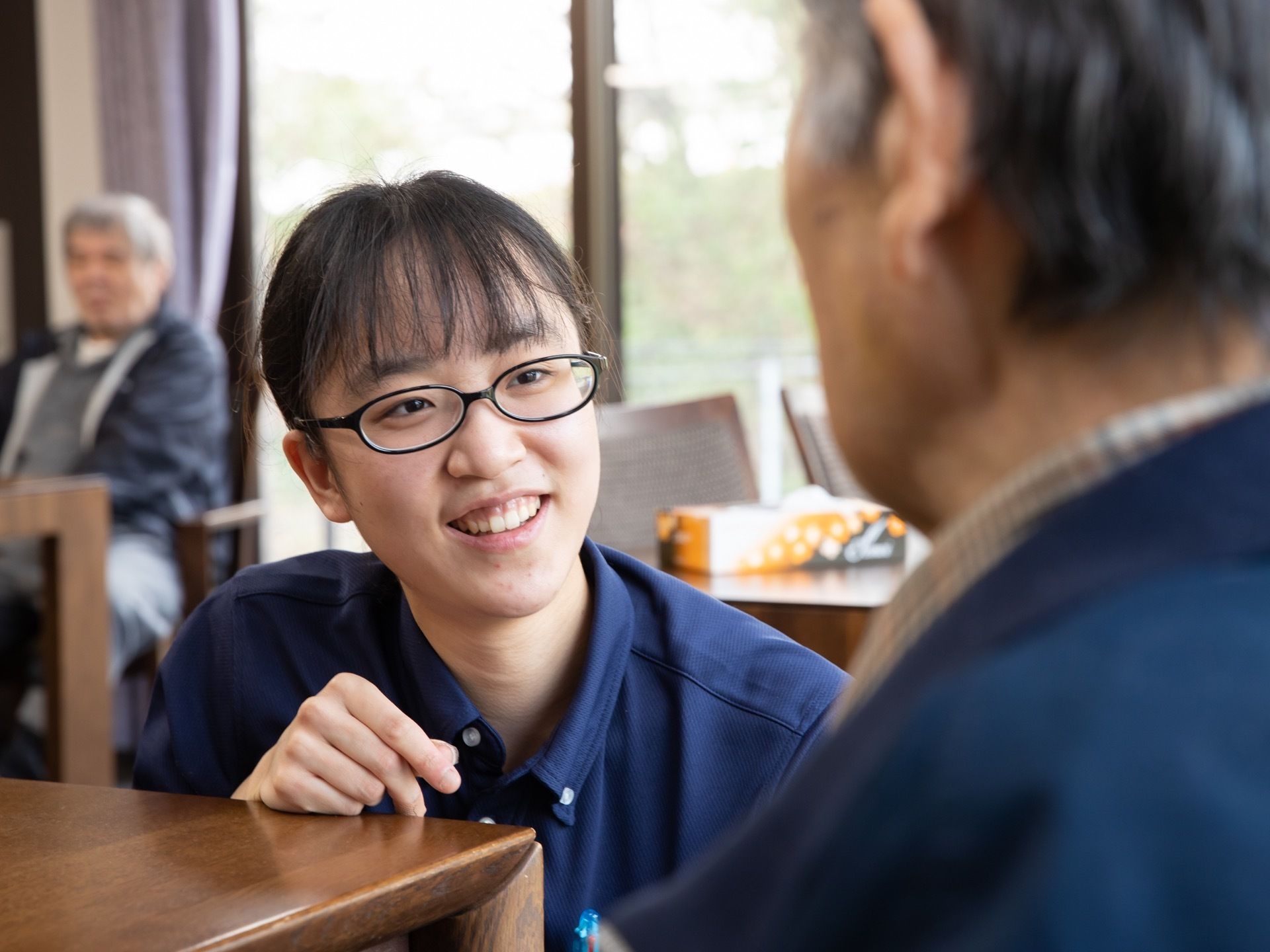 野外の緑が感じられる広い室内。女性スタッフが、男性の傍らで低い姿勢になり、目を見ながら笑顔で話しかけている。