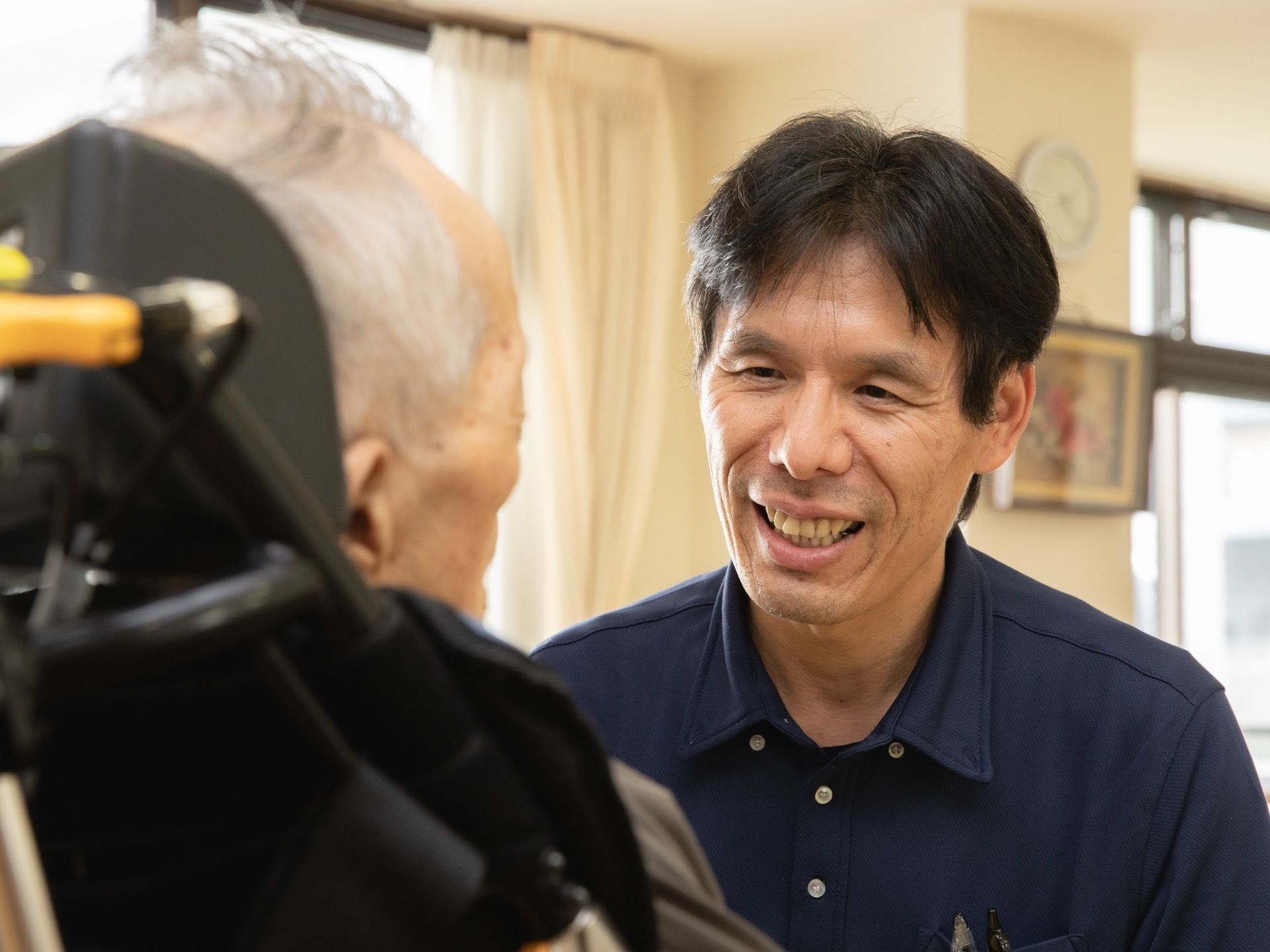 年配の男性スタッフが入居者様に話しかけている様子である。入居者様と同じ目線で優しい対応を取っている。