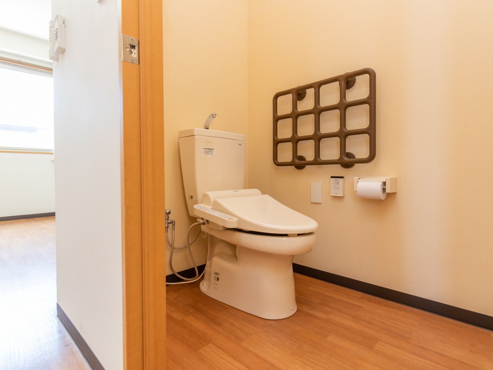 床がフローリングの洋式トイレがある。温水洗浄機能付きで、壁には格子状の茶色い手すりが備えつけられている。