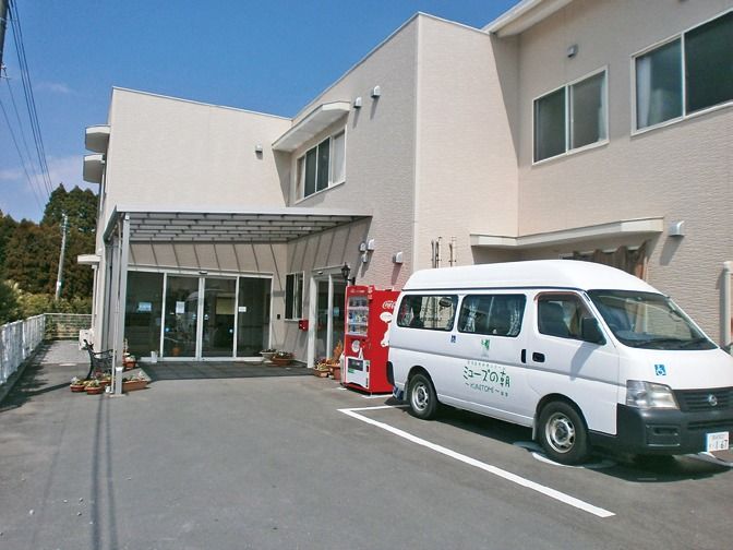 建物の前に施設専用の送迎車が停まっている。出入り口前には屋根があり、自動販売機や植木鉢が置かれている。