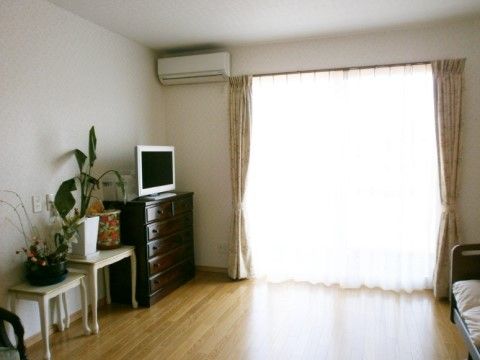 フローリングの床に大きな掃き出し窓のある明るい居室である。エアコンや介護ベッドが備わっている。チェストの上にテレビが置かれている。
