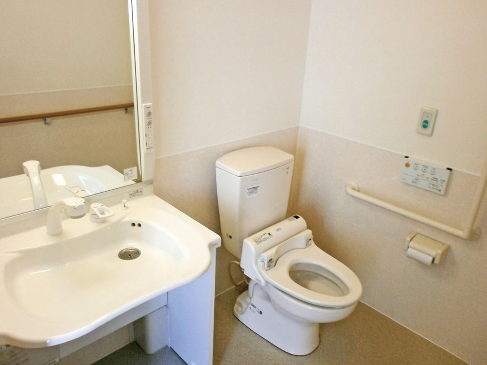 洋式トイレと、車椅子対応型の洗面台の備わった個室である。壁にはナースコールや手すりが設置されている。