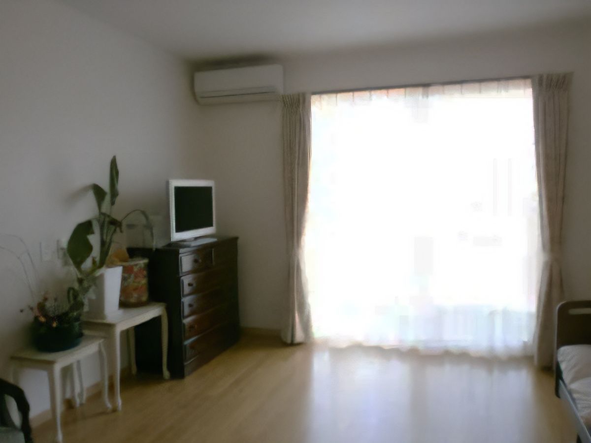 フローリングの床に大きな掃き出し窓のある明るい居室である。エアコンや介護ベッドが備わっている。チェストの上にテレビが置かれている。