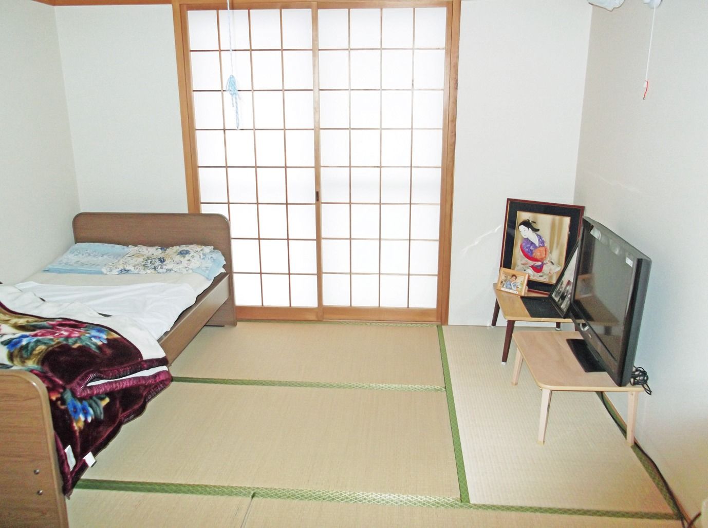 畳敷きの部屋に背の低い木製のベッドが設置されている。壁に沿って小さめの座卓が２台並んでおり、その上にテレビや写真立てが置かれている。