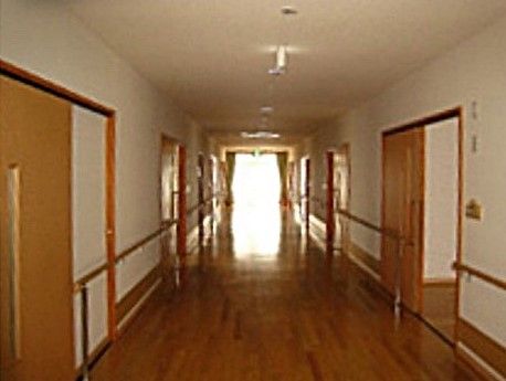 突き当りから外光が入る、明るく幅広い廊下。両側に引き戸タイプの居室扉が並ぶ。壁には床と平行に手すりが付いている。